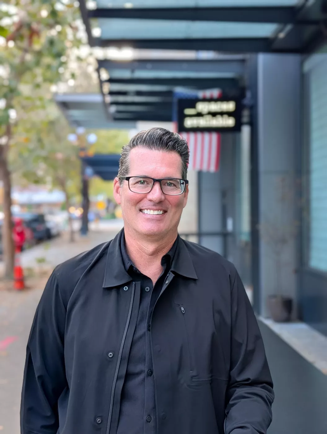 A middle-aged man with short dark hair and glasses smiles at the camera while standing on an urban sidewalk. He is wearing a black button-up jacket over a dark shirt. The background shows a modern building facade with glass and metal elements, trees lining the street, and an out-of-focus sign with an American flag pattern. The overall scene feels casual and friendly in a contemporary city environment.