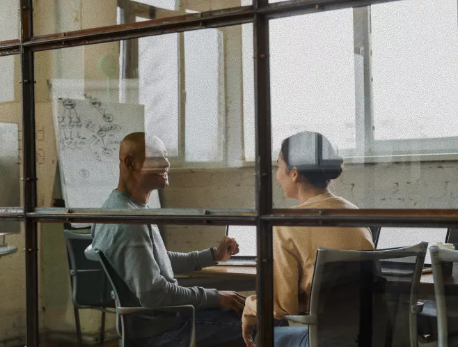 Two colleagues sit facing each other at a desk in a contemporary office space. They appear to be engaged in a relaxed conversation or informal meeting. The scene is viewed through large glass panels, with reflections adding depth to the image. In the background, a whiteboard with sketches and a large window provide a bright, creative atmosphere.