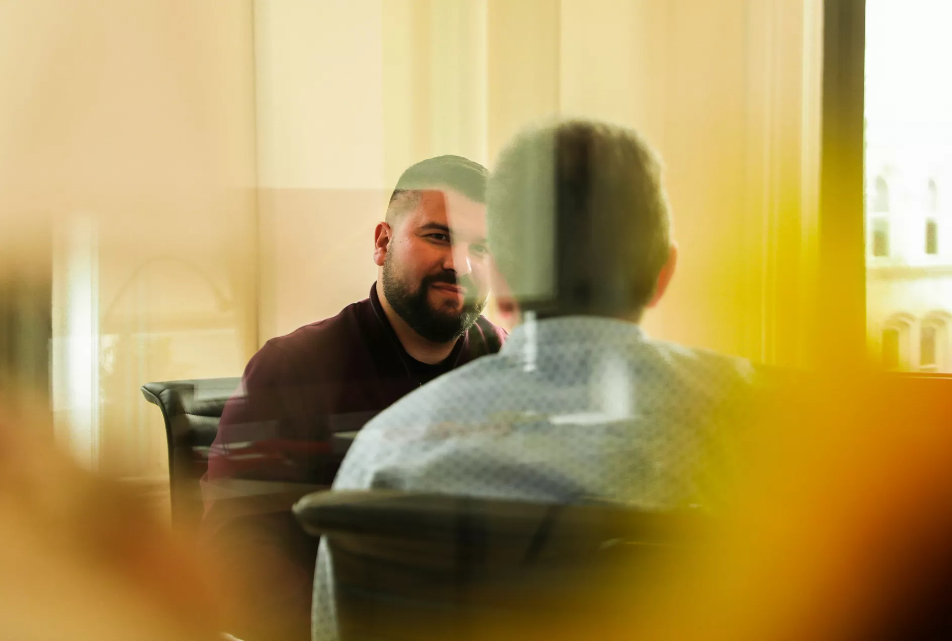 Two men sit facing each other in a modern office setting, engaged in conversation. The scene is viewed through a glass partition, creating soft reflections and abstract yellow and orange blurs in the foreground. One man is seen from behind while the other faces the camera, suggesting a meeting or one-on-one discussion. Natural light filters into the room, giving the image a warm and relaxed atmosphere.