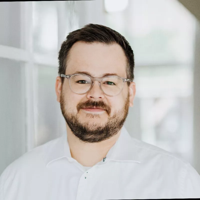 A man wearing a crisp white button-up shirt is pictured indoors, standing next to a window with natural light streaming in. The background is softly blurred, suggesting a modern, bright interior setting. The subject appears to be an adult male with short brown hair and a beard. No visible text, logos, or numeric values are present in the image.