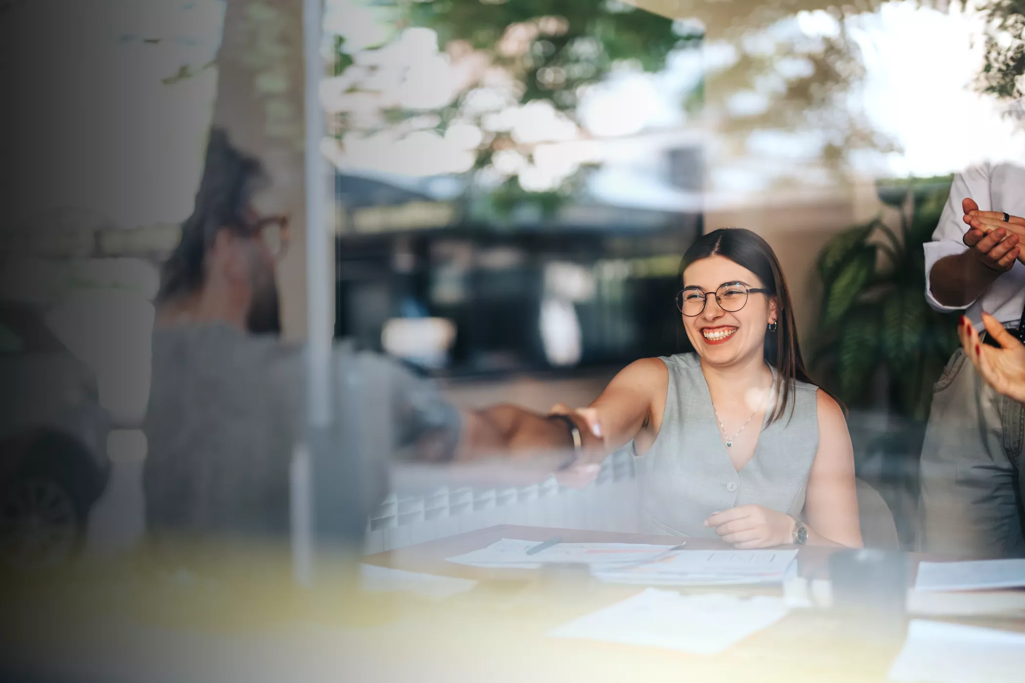 A woman sits at a table in a contemporary office environment, reaching out to shake hands with another person across the table. Papers and documents are spread out in front of her, suggesting a professional discussion or negotiation. The scene is bright, with natural light and greenery visible through large windows, creating a collaborative and welcoming atmosphere. Another person stands nearby, clapping or gesturing in support.