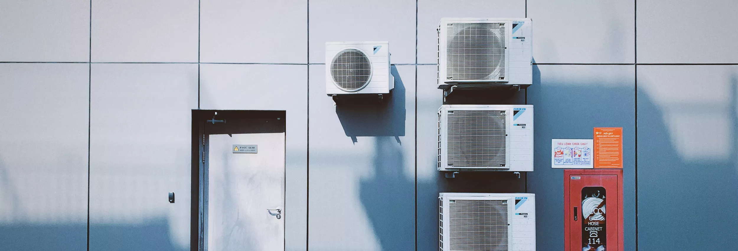 The image shows several wall-mounted air conditioning units installed on the exterior of a modern building. A metal door and a red utility box are also visible on the paneled facade. The scene is evenly lit, highlighting the clean, geometric lines of the structure. Subtle shadows from the units and fixtures add depth to the otherwise minimal industrial setting.