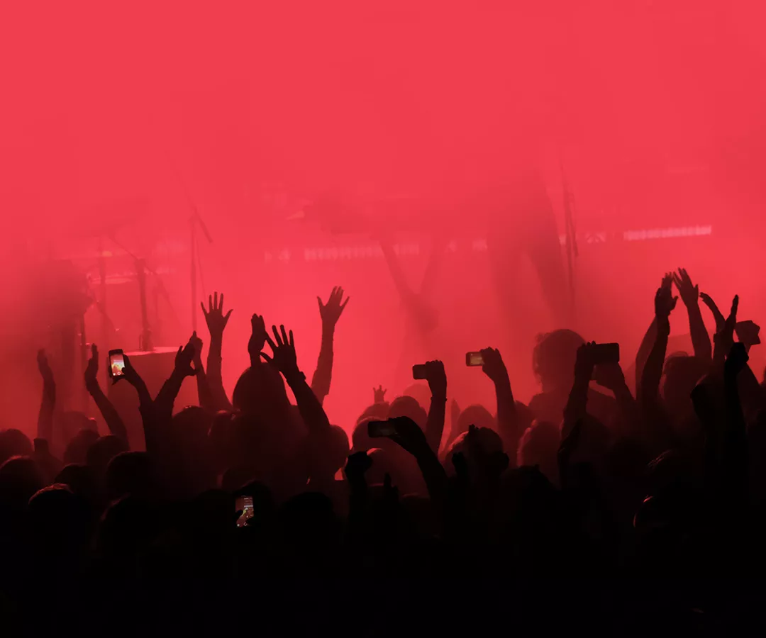 Red background with a crowd of cheering people at a concert. People with their hands up