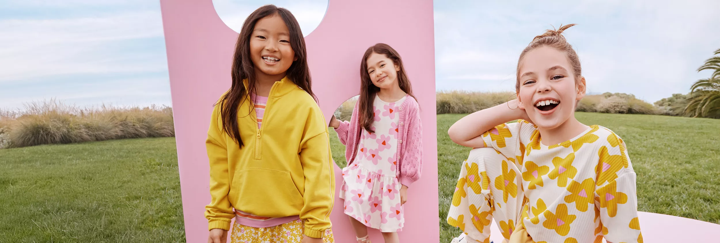 Three young girls are pictured outdoors, standing and sitting in front of a large pink geometric backdrop. They are dressed in bright, patterned clothing featuring yellow and pink hues, with playful prints and casual styles. The grassy field and blue sky create a cheerful, relaxed atmosphere, emphasizing spring or summer fashion for children. No visible text or numbers are present in the image.