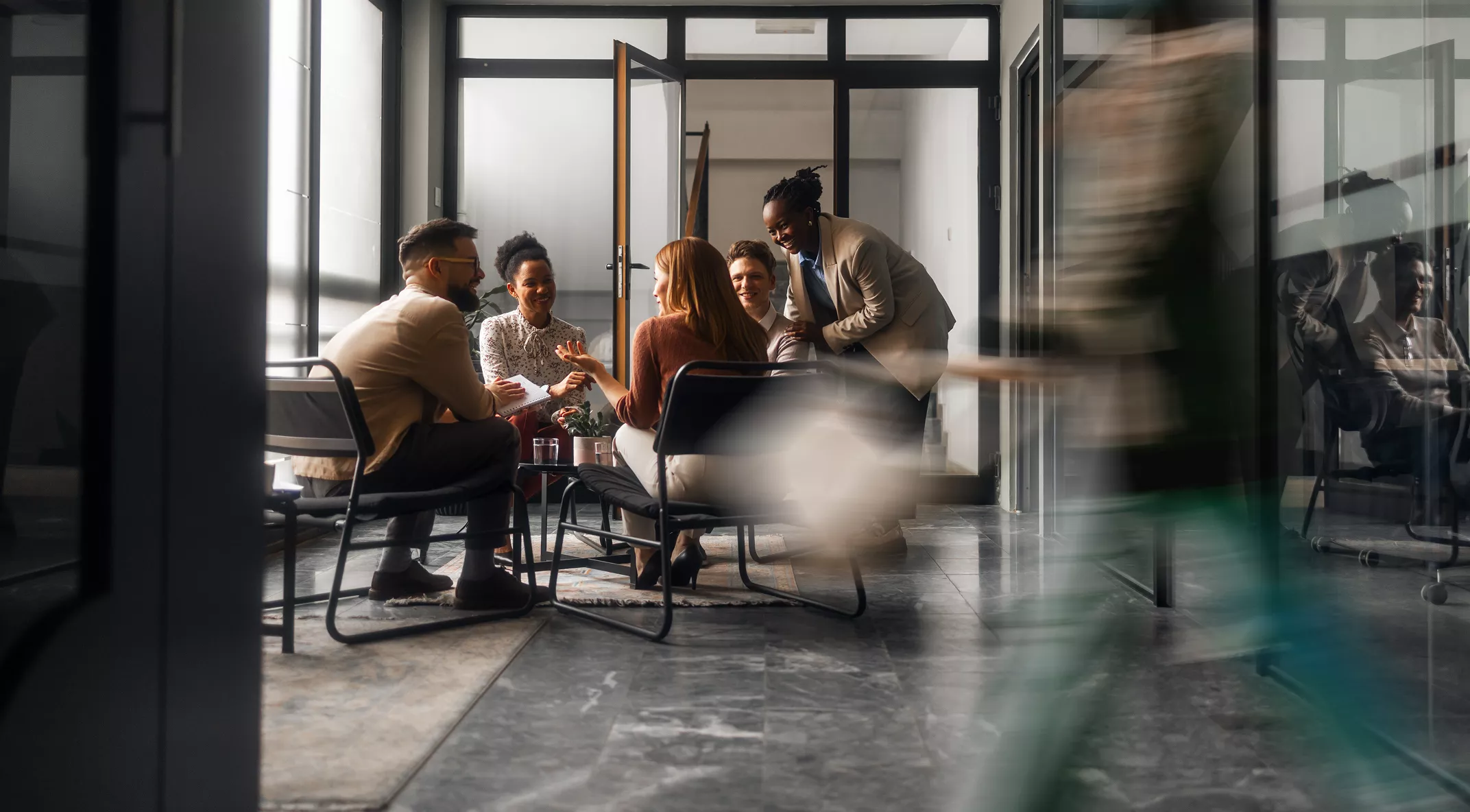 A small group of colleagues sit together in a glass-walled meeting room, engaged in a collaborative discussion. One person stands and leans over the table while others listen and smile, suggesting a relaxed yet productive atmosphere. The office features contemporary furniture, large windows, and polished concrete floors. A blurred figure walks past in the foreground, adding a sense of motion and busy workplace energy.