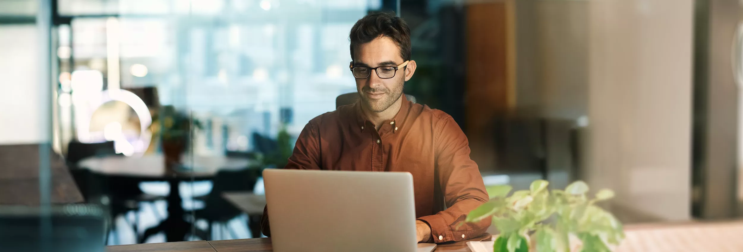 A person sits at a desk working on a laptop in a contemporary office environment. The individual wears a long-sleeve shirt and is positioned behind a glass partition, suggesting a meeting room or quiet workspace. Soft, warm lighting and blurred office furniture in the background create a modern, professional atmosphere. A green plant in the foreground adds a touch of nature to the otherwise sleek interior.