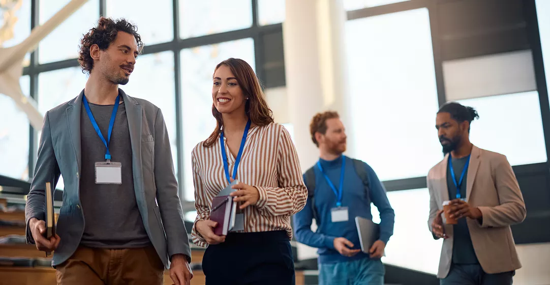 A group of four adults, each wearing a blue lanyard with a visible badge, walk together inside a bright, modern office building. The setting features large windows and a staircase, suggesting a collaborative or conference environment. The individuals are dressed in business casual attire and carry notebooks or folders. The overall mood is professional and dynamic, with natural daylight illuminating the scene.