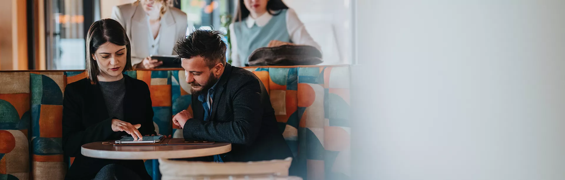 Two adults are seated at a patterned booth table, engaged in a discussion with documents or a notebook. In the background, two more adults stand, one holding a tablet, suggesting collaboration or presentation. The setting is a contemporary cafe or casual workspace with geometric upholstery and warm lighting. The overall mood is professional and focused, with a mix of business casual attire. No visible text or numbers are present in the image.