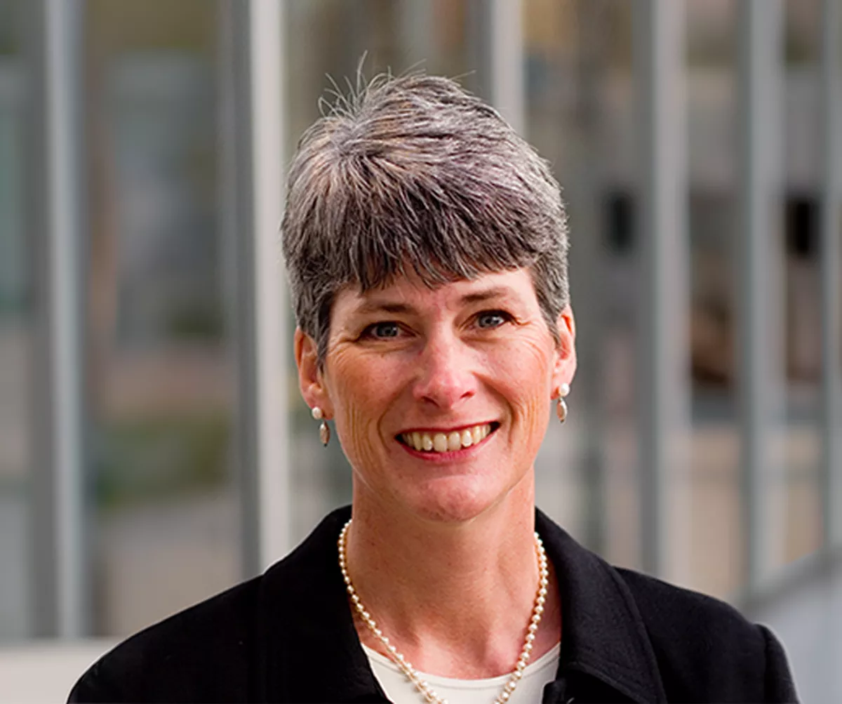 A professional woman is shown in a close-up portrait with her face intentionally blurred for privacy. She has short gray hair and wears a dark blazer over a light top. A pearl necklace and small earrings add a formal touch to her appearance. The background features an office-like setting with vertical architectural elements softly out of focus.