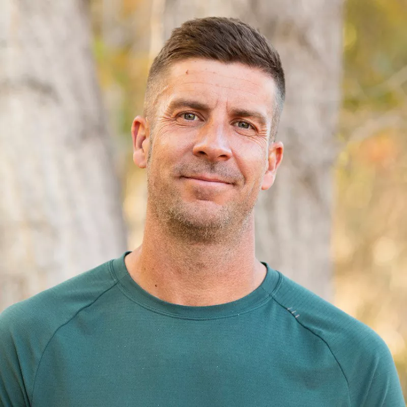 Portrait of an adult man standing outdoors, looking directly at the camera with a slight smile. He wears a long-sleeve green athletic-style shirt, and the background shows softly blurred trees and warm natural light. The image has a relaxed, approachable mood suitable for lifestyle or outdoor themes. Neutral tones in the background contrast with the solid color of his shirt, keeping focus on his face.