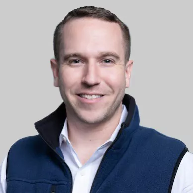 A man is pictured wearing a blue fleece vest over a white collared shirt against a plain grey background. The subject's face is obscured, focusing attention on the clothing and overall style. The setting is neutral, with no visible text, numbers, or identifiable brands. The image conveys a casual yet professional mood.