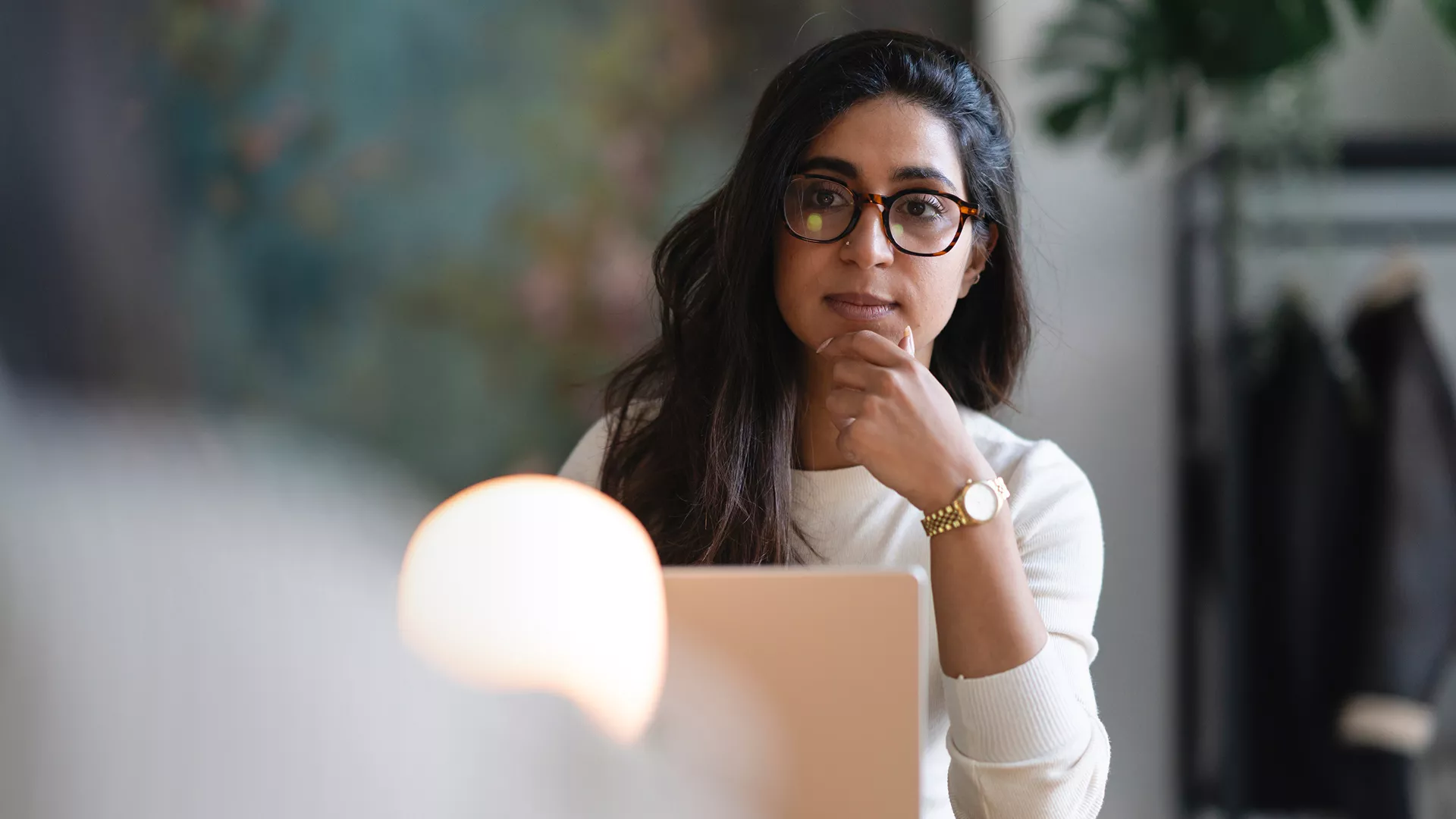 A woman with long dark hair sits at a desk, working on a laptop in a contemporary office environment. She wears a light sweater and a gold wristwatch, with her hand resting near her face. The background features a blurred plant and abstract wall art, creating a calm and professional atmosphere. Soft lighting adds warmth to the scene.