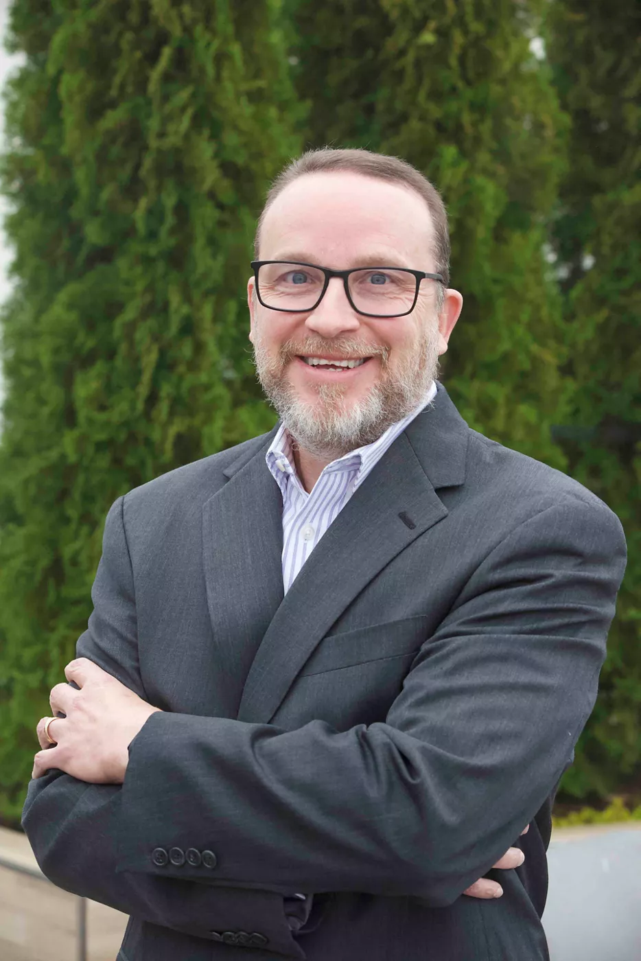 A middle-aged man in a dark suit jacket and striped dress shirt stands outdoors with his arms crossed. He is smiling confidently, wearing glasses and a neatly trimmed beard. Tall green trees form a natural background, suggesting an outdoor or corporate campus environment. The overall scene conveys professionalism and approachability.