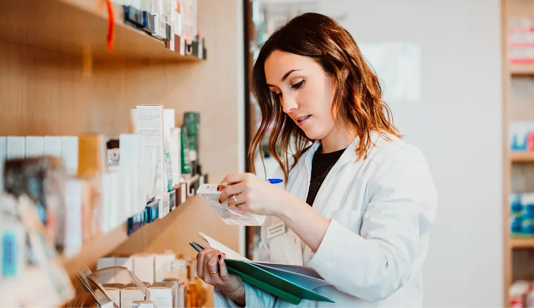 Female pharmacist stnading in front of shelf looking at medicine while holding book