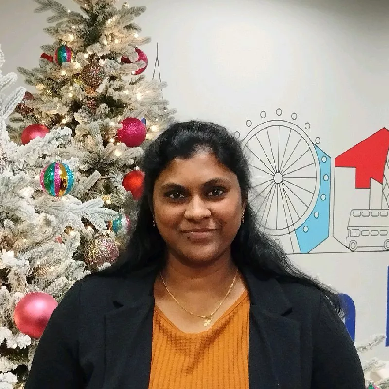 A woman stands in front of a frosted Christmas tree adorned with colorful ornaments and red baubles. The setting appears to be an office space, featuring a wall mural with a Ferris wheel and cityscape elements. The mood is cheerful and festive, with a warm palette of gold, red, and green. No visible text or numeric values are present in the image.