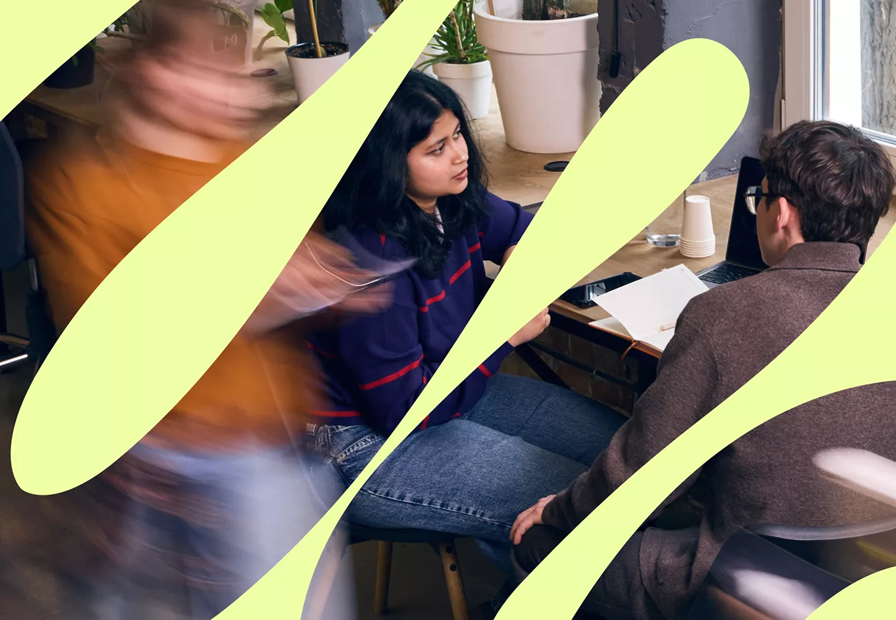 Two coworkers sit at a shared desk in a modern office, engaged in conversation. A notebook, pen, and laptop are on the table, suggesting a casual meeting or brainstorming session. Blurred motion of a person walking by adds energy and a sense of movement to the scene. Bright abstract yellow shapes overlay the image, giving it a contemporary, artistic feel.