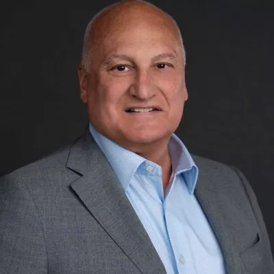 Portrait of a business professional wearing a gray suit jacket over a light blue dress shirt. The subject is posed against a plain dark background, suggesting a formal studio or corporate setting. The image focuses on the upper body and attire, with the face obscured. The overall style is clean, professional, and suitable for corporate or business-related use.