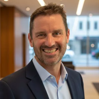 A middle-aged man in a business suit smiles warmly at the camera in a modern office lobby. The background shows large windows and contemporary interior elements with soft, even lighting. His open, friendly expression and relaxed posture convey approachability and confidence. The image suggests a corporate or professional environment suitable for business or executive profiles.