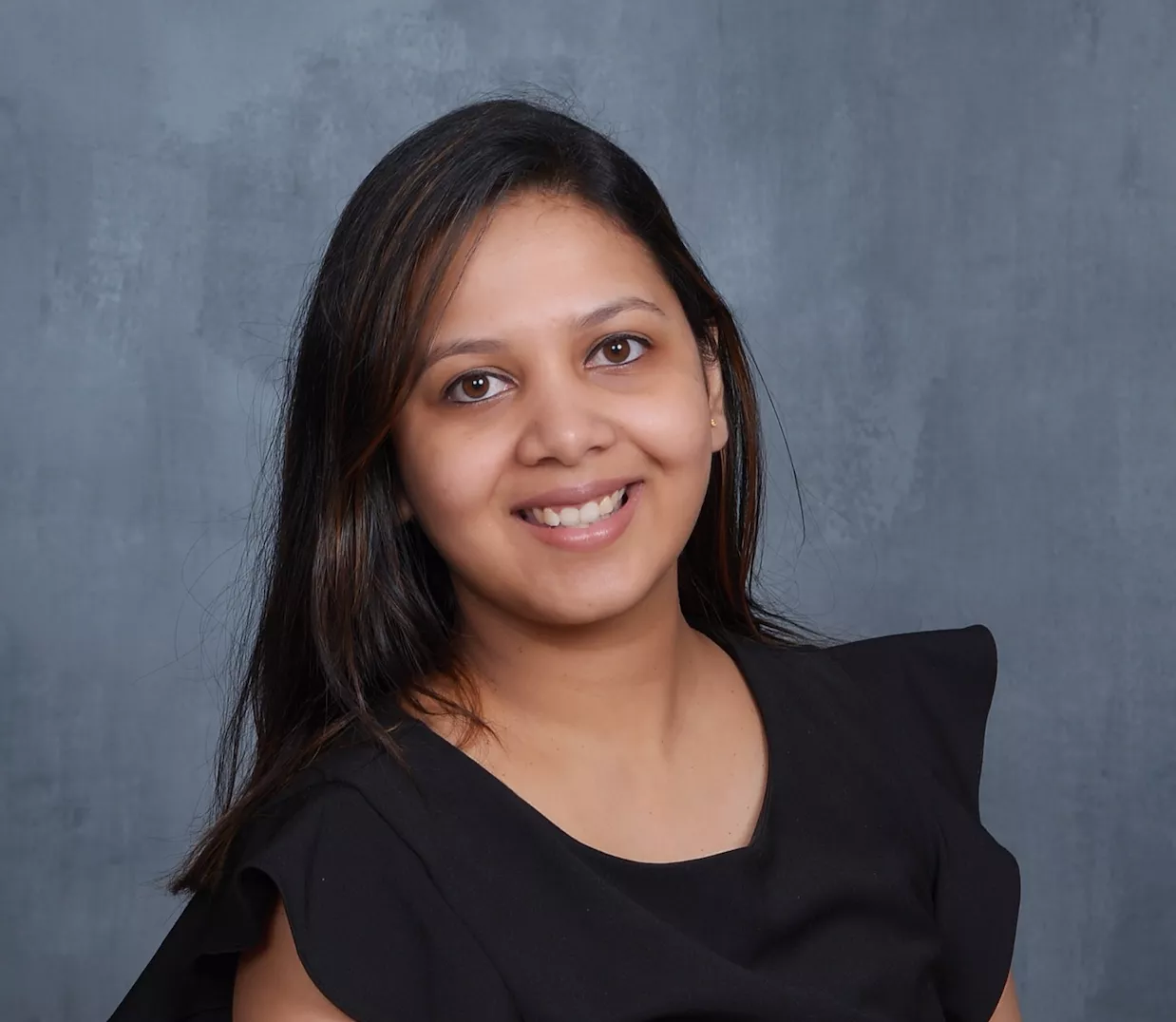 A young woman smiles warmly in a studio portrait. She is wearing a black top with ruffled sleeves and has straight, shoulder-length hair. The background is a neutral gray with a subtle textured pattern. Soft lighting highlights her face, creating a friendly and approachable appearance.