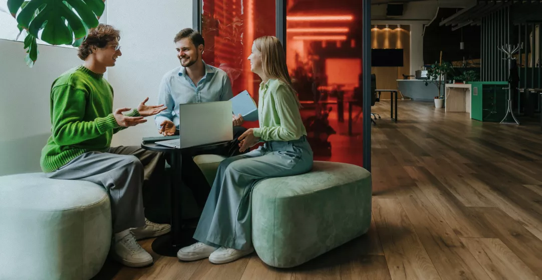 Three coworkers sit together around a small round table in a contemporary office space. A laptop is open between them as they engage in a friendly discussion, suggesting teamwork and collaboration. The setting features wooden floors, large plants, soft green seating, and red glass partitions that add a colorful, modern touch. The overall atmosphere feels casual, creative, and welcoming.