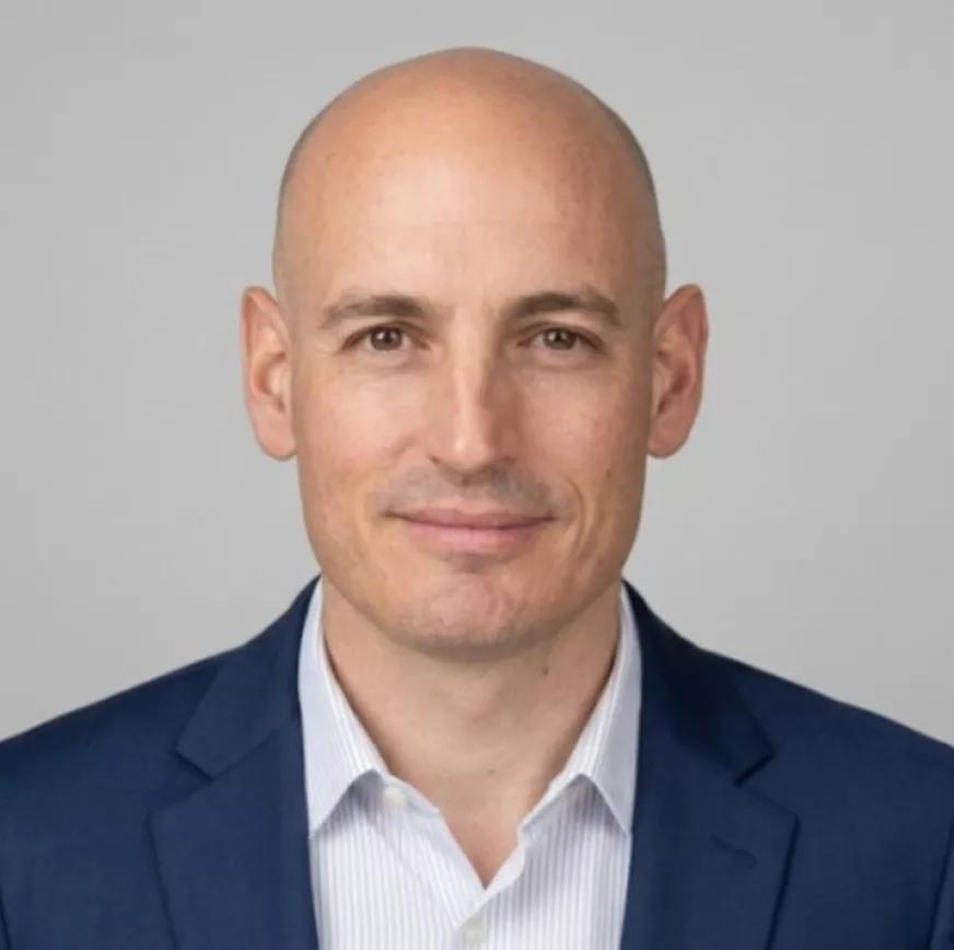 Head-and-shoulders portrait of a bald man wearing a blue suit jacket and a light striped dress shirt. He is facing the camera with a neutral, confident expression against a plain light gray background. The image has a clean, corporate style suitable for business or professional profiles.
