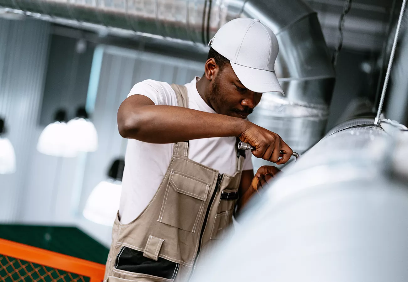 A technician in work overalls inspects a large metal ventilation duct in an industrial setting. He wears a white cap and focuses on adjusting or repairing a section of the ductwork. The environment features exposed pipes, ducts, and bright overhead lighting, suggesting a factory or commercial facility. The image emphasizes hands-on maintenance, technical skill, and industrial infrastructure.