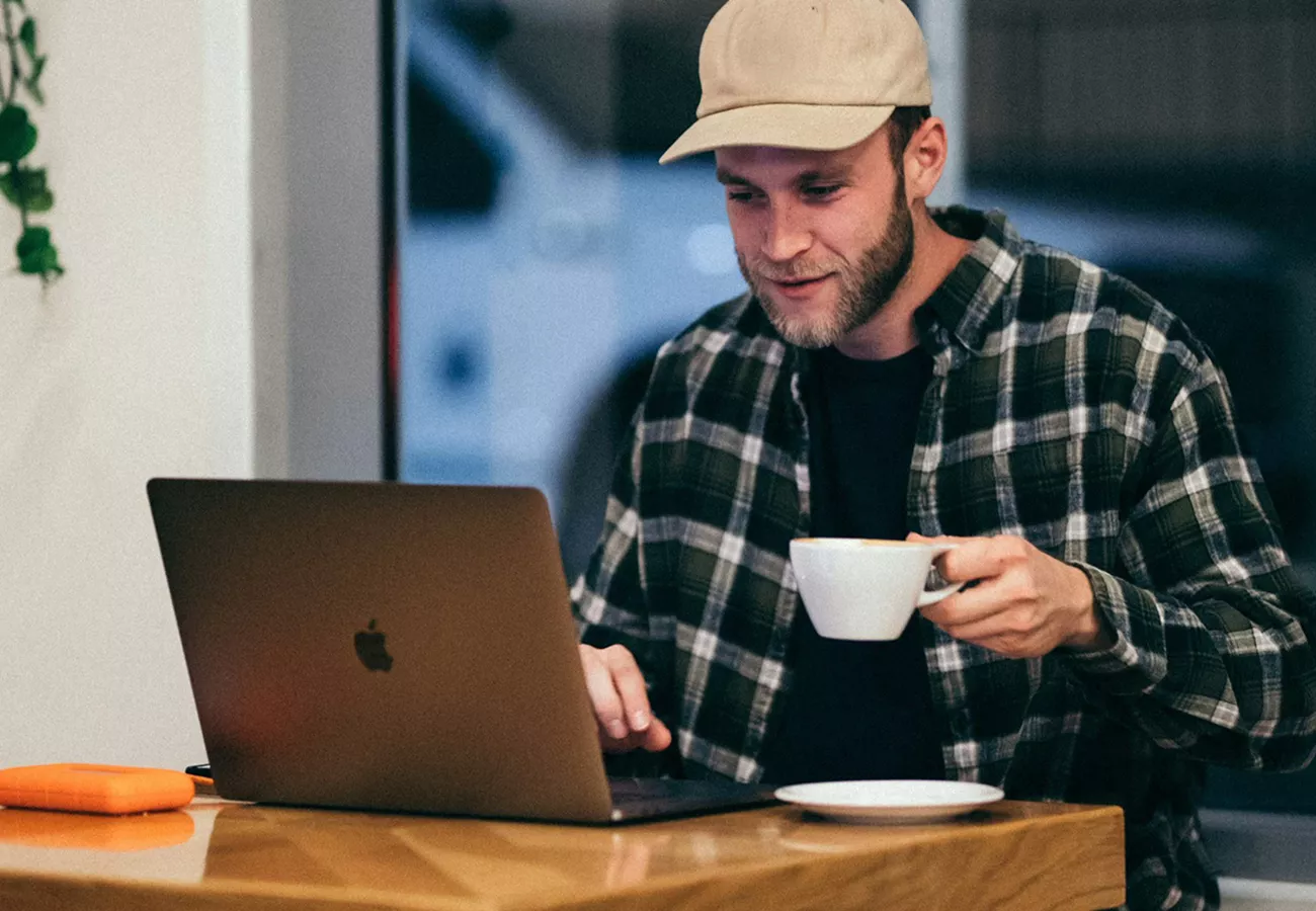 A person sits at a wooden table working on a laptop while holding a white coffee cup. The setting appears to be a cozy cafe or casual workspace with a window in the background. The individual wears a plaid shirt and a light-colored cap, suggesting a relaxed, informal atmosphere. A small orange external drive or accessory lies on the table beside the laptop, adding a pop of color to the scene.
