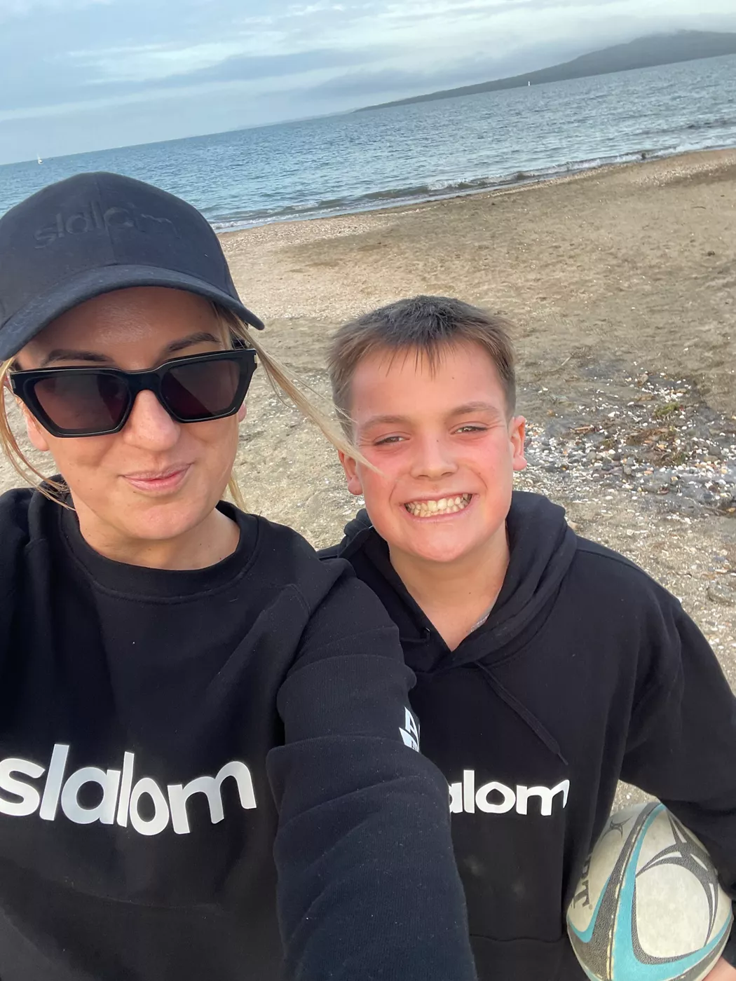 A woman and a young boy smile at the camera while standing on a sandy beach near the water. Both wear black hoodies with the word "slalom" clearly visible on the front, and the woman also wears a black cap and sunglasses. The boy holds a ball under his arm, suggesting casual playtime by the shore. The background shows calm sea water and distant land under a cloudy sky, creating a relaxed coastal atmosphere.