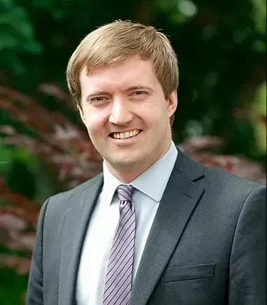 A man wearing a dark suit and a light-colored, striped tie stands outdoors. The background features lush green foliage, suggesting a garden or park setting. The man's face is intentionally blurred for privacy. The overall mood is formal and professional, with natural lighting.