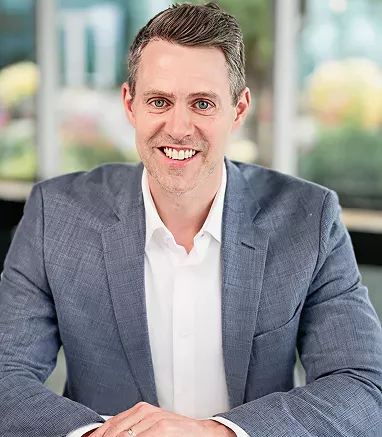 A man in a blue checkered suit jacket and white dress shirt is seated at a table in an outdoor or glass-walled setting. The background features blurred greenery and modern architecture, suggesting a professional or corporate environment. The subject's face is obscured, focusing attention on the attire and posture. The overall mood is formal and businesslike, with natural daylight illuminating the scene.