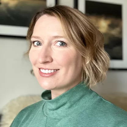 A woman with short, wavy blonde hair smiles at the camera while wearing a light green turtleneck top. She is indoors, seated in front of framed artwork on a neutral-colored wall. Soft, natural lighting highlights her face and creates a warm, welcoming atmosphere. The overall scene feels casual and contemporary, suitable for professional or lifestyle contexts.