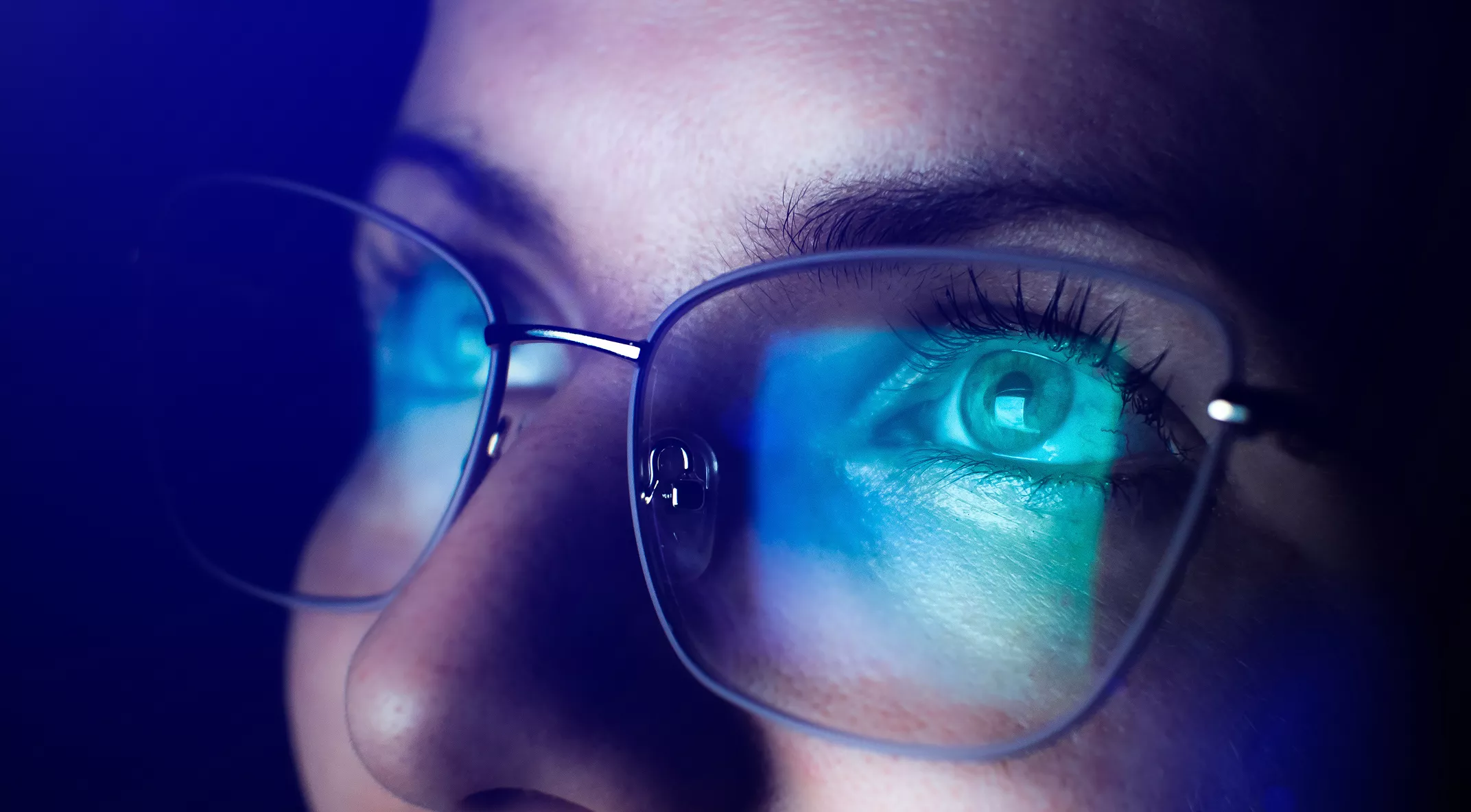 A close-up image shows a woman's face with clear eyeglasses reflecting a blue light screen. The setting suggests she is looking at a digital device, possibly a computer or smartphone. The visuals highlight the blue glow on her lenses, emphasizing eye protection from digital screens. The overall mood is focused and modern, with cool blue tones.