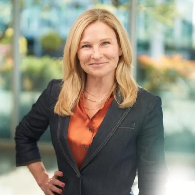 A confident woman stands indoors, smiling directly at the camera. She is dressed in a dark blazer over an orange blouse, suggesting a professional or business setting. The background shows a bright, modern office or corporate environment with large windows and soft natural light. The overall scene conveys approachability, leadership, and professionalism.