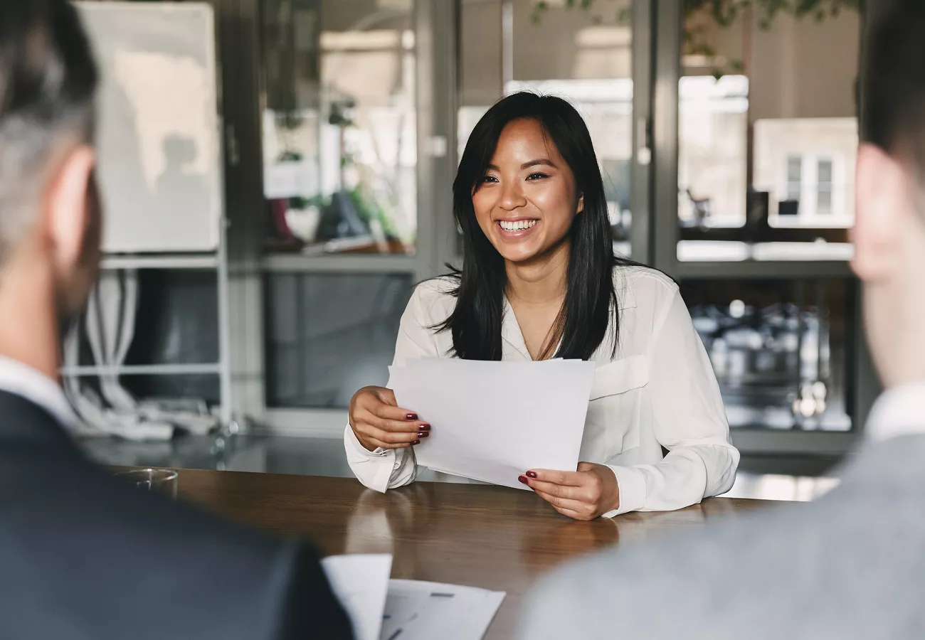 A woman sits at a conference table holding papers, facing two people across from her. The scene takes place in a contemporary office with glass walls and visible greenery outside. The atmosphere is professional and focused, with neutral tones and natural light. No visible text or numbers are present in the image.