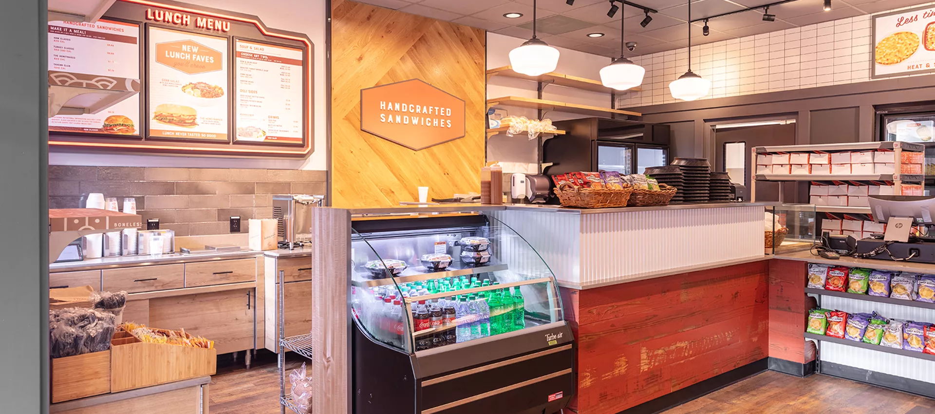 A clean and inviting sandwich shop interior features a refrigerated display case with bottled drinks and a variety of snacks on shelves. Prominent menu boards display lunch options and prices, with clear signage reading 'Handcrafted Sandwiches.' The setting is well-lit with warm wood accents and a casual, welcoming atmosphere. No people are present, and the space is organized for quick service.