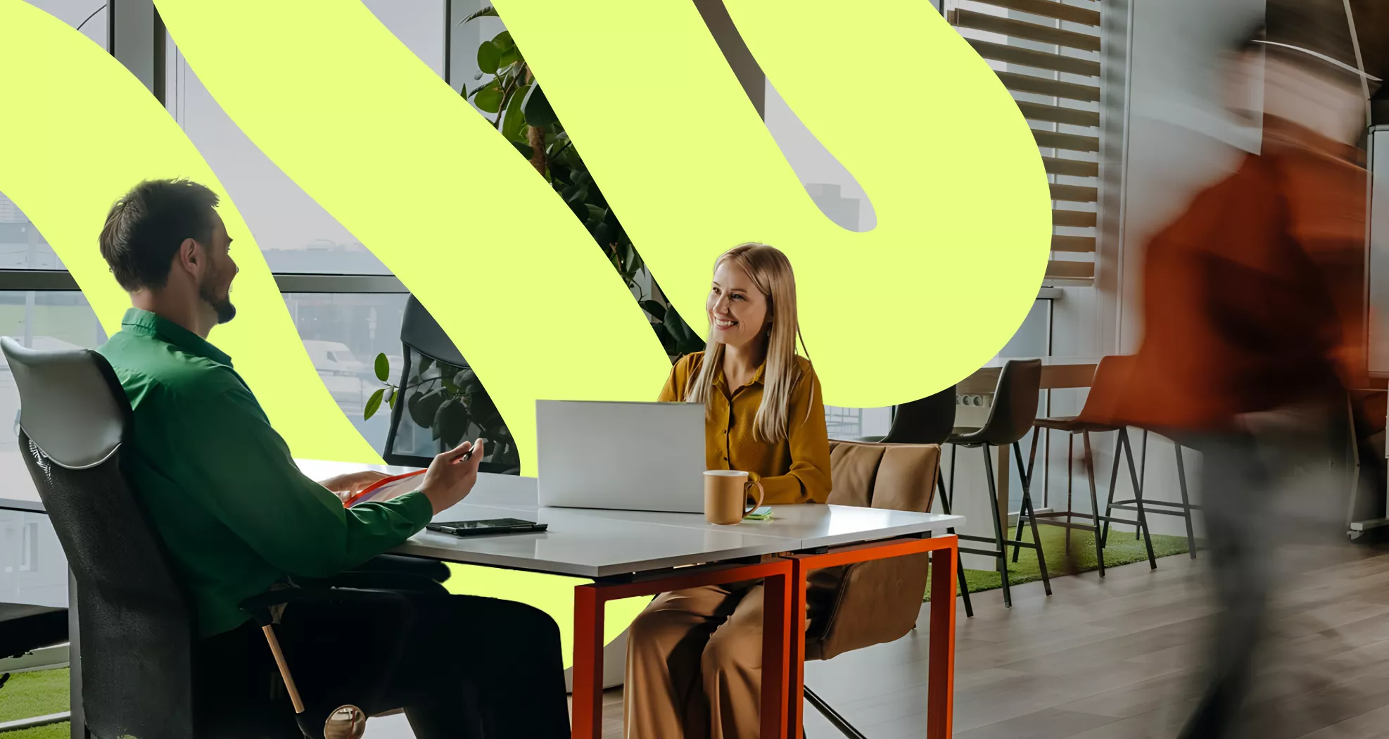 Two colleagues sit across from each other at a desk in a bright, contemporary office space. The woman works on a laptop while the man faces her, suggesting a casual meeting or discussion. A blurred person walks past in the foreground, adding a sense of motion and activity. Large abstract yellow shapes and floor-to-ceiling windows create a modern, creative atmosphere.
