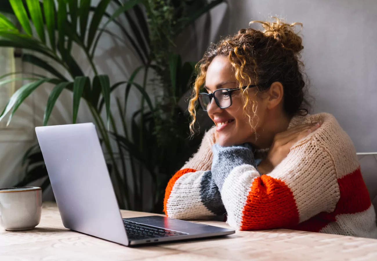 A woman sits at a wooden table with a laptop open in front of her, appearing focused and thoughtful. The setting is a cozy indoor space with lush green plants in the background, suggesting a home office or relaxed workspace. She wears a colorful striped sweater with red, orange, and gray tones, adding warmth to the scene. The overall mood is calm and introspective, with natural light illuminating the area.