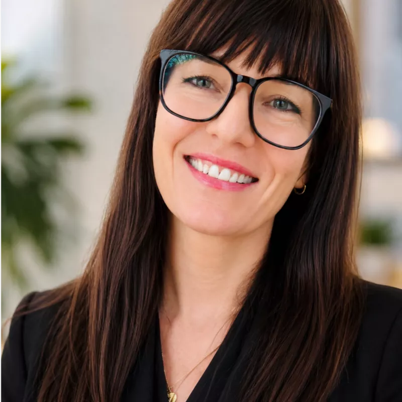 Portrait of a smiling woman with long dark hair and black eyeglasses. She is dressed in a dark blazer, suggesting a professional or business setting. The background is softly blurred with hints of indoor plants and light, creating a bright and welcoming atmosphere. Her friendly expression and direct gaze convey confidence and approachability.
