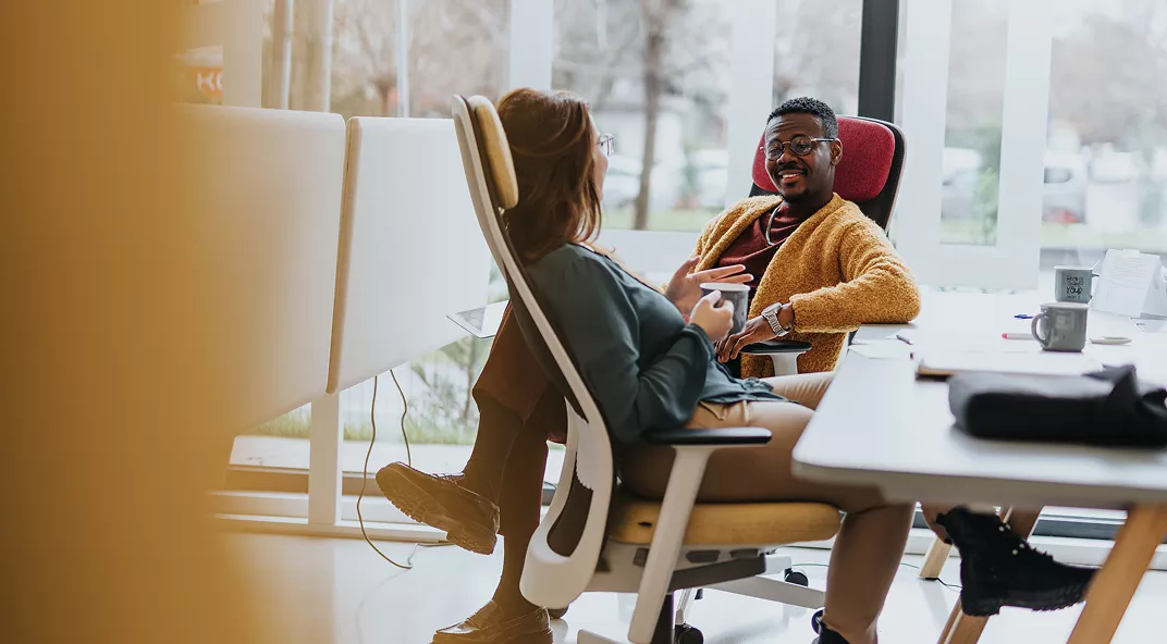 Two people are seated in ergonomic office chairs, engaged in a relaxed conversation in a contemporary workspace. The setting features large windows, natural light, and a casual atmosphere. Both individuals appear to be young adults, one male and one female, dressed in comfortable business casual attire. The workspace includes desks, a laptop, and a coffee cup, contributing to a collaborative and informal mood.