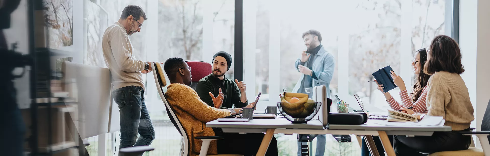 A diverse group of young adults is engaged in a collaborative meeting around a large table in a bright, modern office. Some team members are seated and actively discussing ideas, while others stand near large windows, contributing to the conversation. Laptops, notebooks, and a bowl of fruit are visible on the table, emphasizing a casual yet productive atmosphere. The setting features natural light and a contemporary interior design.