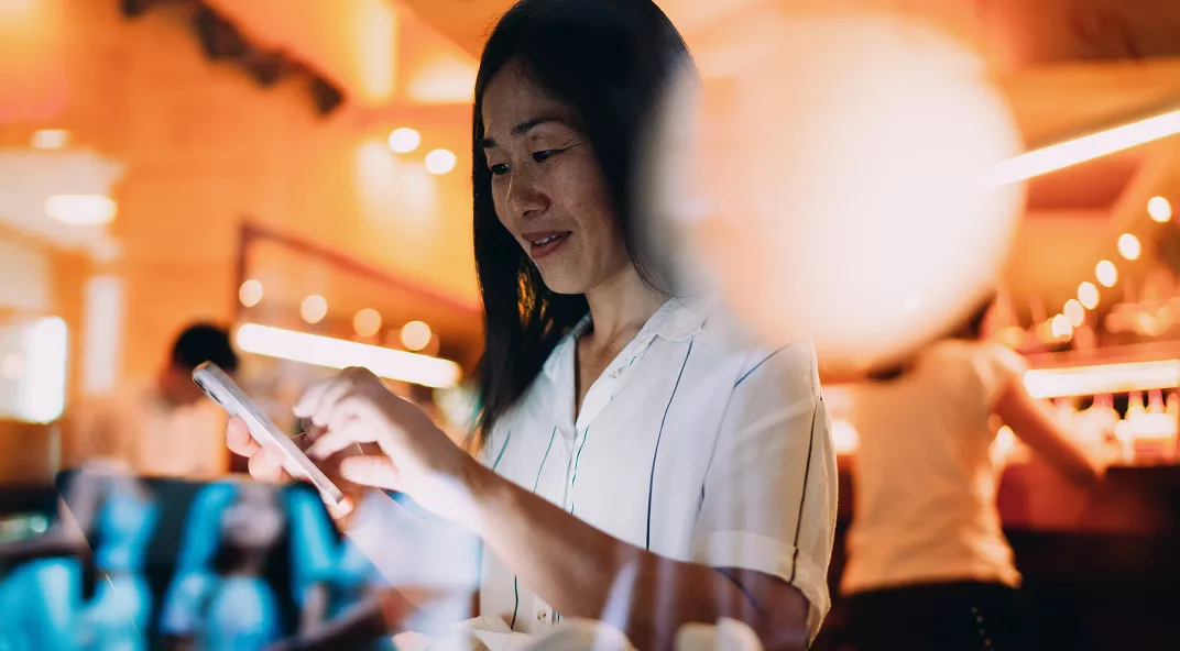 A young woman is focused on her smartphone while seated in a contemporary cafe. The background features warm lighting and blurred patrons, creating a lively yet cozy atmosphere. She wears a light-colored blouse, and the overall palette is dominated by amber and soft tones. The scene conveys a sense of casual social interaction and modern lifestyle.