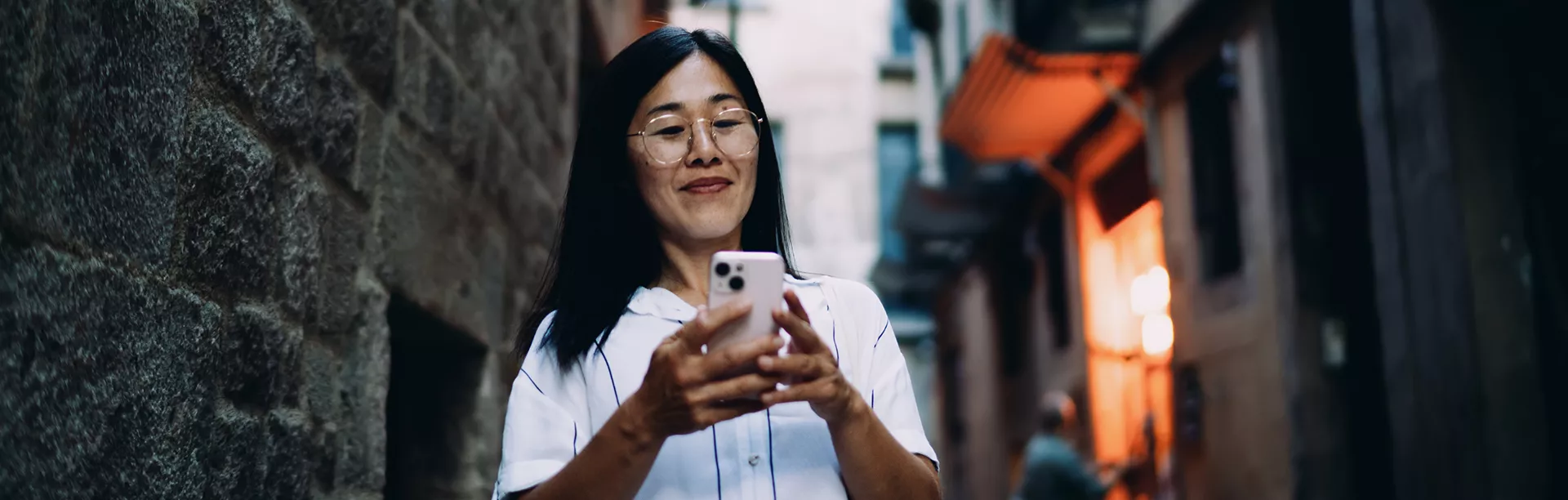 A woman stands in a narrow urban alley, holding and using a smartphone. She is dressed in a white shirt, and the background features textured stone walls and blurred city elements. The lighting is natural, creating a casual and candid atmosphere. No visible text or numbers are present in the image.