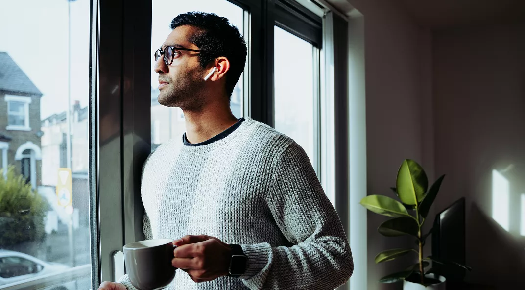 A young adult male stands indoors by a large window, holding a white mug in one hand. He is wearing a textured white sweater and a smartwatch, gazing outside with natural light illuminating the scene. The background includes a leafy green plant and a glimpse of residential buildings through the window. The overall mood is calm and contemplative, with a modern, cozy interior setting.