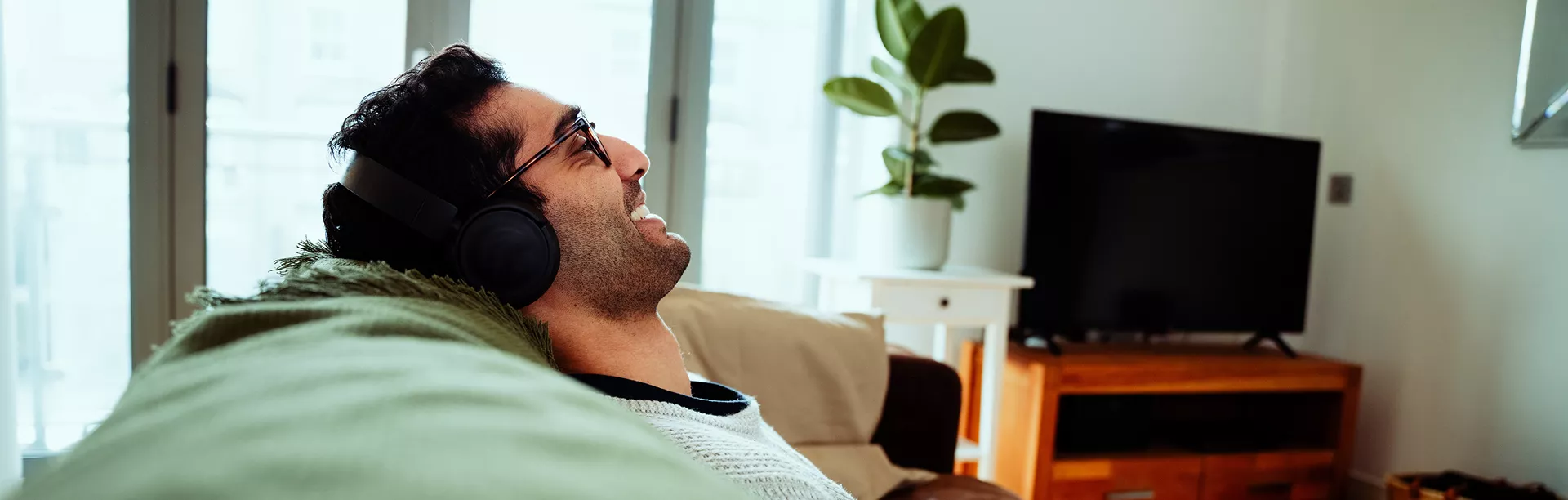 A man is seen reclining on a couch, wearing headphones and facing a television in a bright, modern living room. The space features a large window, a wooden TV stand, and a potted plant, creating a cozy and contemporary atmosphere. The overall mood is relaxed and casual, with natural light filling the room. No visible text or numbers are present in the image.
