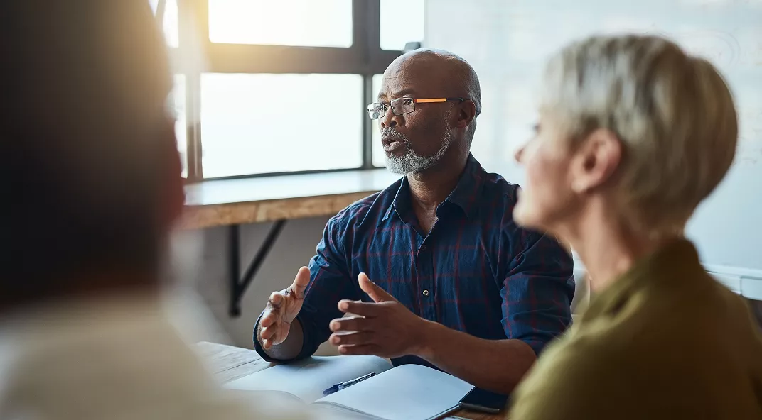A group of adults is seated around a table in a modern office environment, actively participating in a discussion. The main subject, a middle-aged man, gestures while speaking, suggesting a collaborative or brainstorming session. Natural light streams through large windows, creating a bright and focused atmosphere. Notebooks and documents are visible on the table, emphasizing a business or team setting.