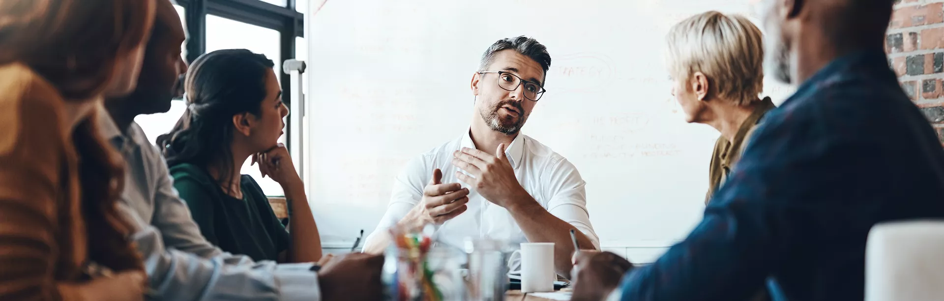 A group of adults sits around a conference table, actively participating in a business meeting. The setting is a modern office with a whiteboard in the background, featuring handwritten notes and diagrams. The atmosphere appears focused and collaborative, with natural light illuminating the workspace. No visible text or numbers are clearly identifiable in the image.