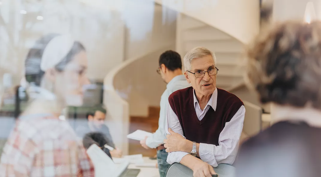 A group of people are engaged in conversation in a contemporary office setting. The scene features a prominent spiral staircase in the background and glass walls, creating an open and airy atmosphere. The individuals appear to be of mixed ages and genders, with one older man in focus wearing a maroon sweater vest. The mood is collaborative and professional, with neutral and warm tones throughout.
