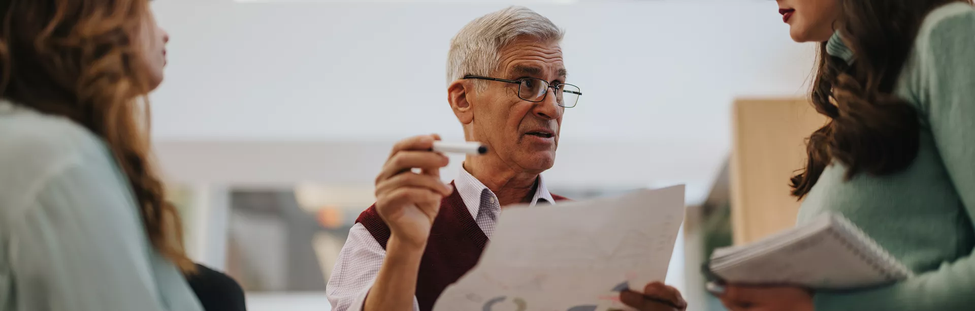 An older man is holding a pen and a document, engaging in conversation with two women in a modern indoor setting. The group appears to be discussing paperwork or a project, with one woman holding a notebook. The atmosphere is professional and collaborative, featuring neutral tones and soft lighting.