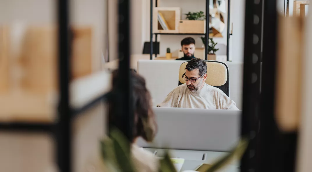 The image shows a contemporary office environment with people working at desks. The setting features open shelving, neutral tones, and modern furniture. Two individuals are visible, one in the foreground and another further back, both seated and focused on their tasks. The atmosphere appears collaborative and professional, with natural light and organized decor.