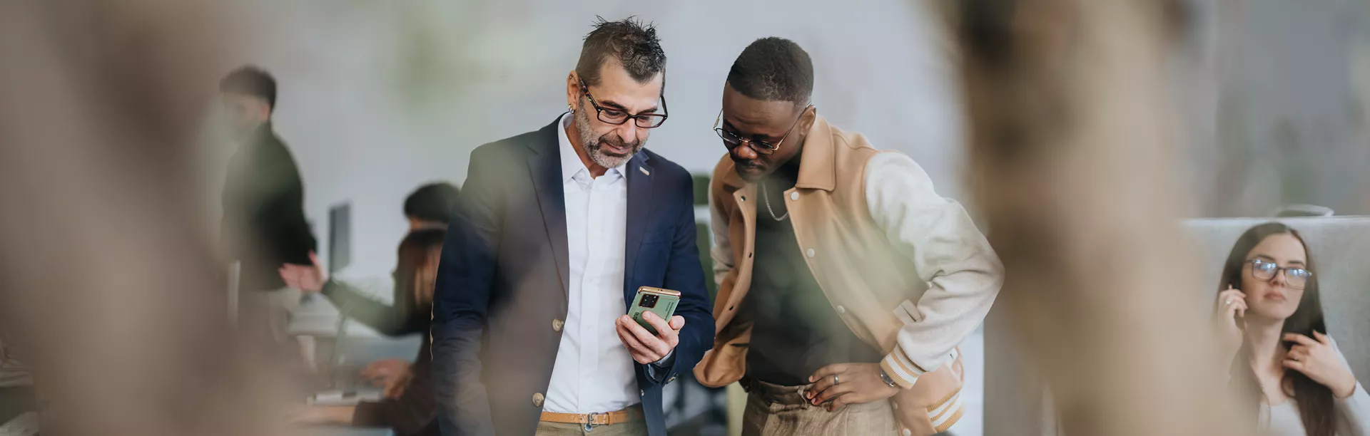 Two adults are engaged in conversation while looking at a smartphone in a contemporary office environment. The scene includes blurred foreground elements and other people working in the background, creating a collaborative atmosphere. The individuals appear focused and professionally dressed, contributing to a productive and dynamic workspace. No visible text or numbers are present in the image.